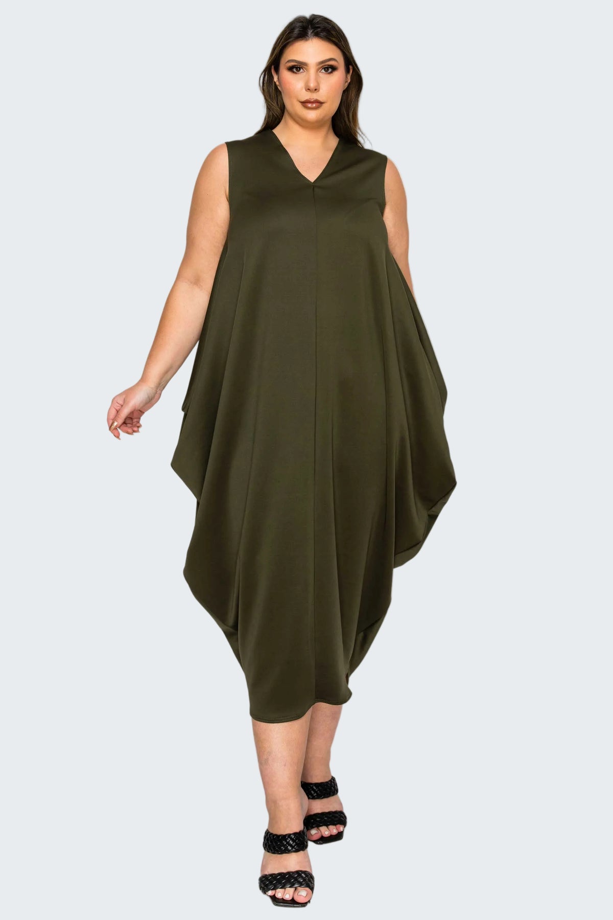 A woman modeling a sleeveless olive green V-neck dress with a draped, asymmetrical hem, paired with black strappy heels, against a plain white background.