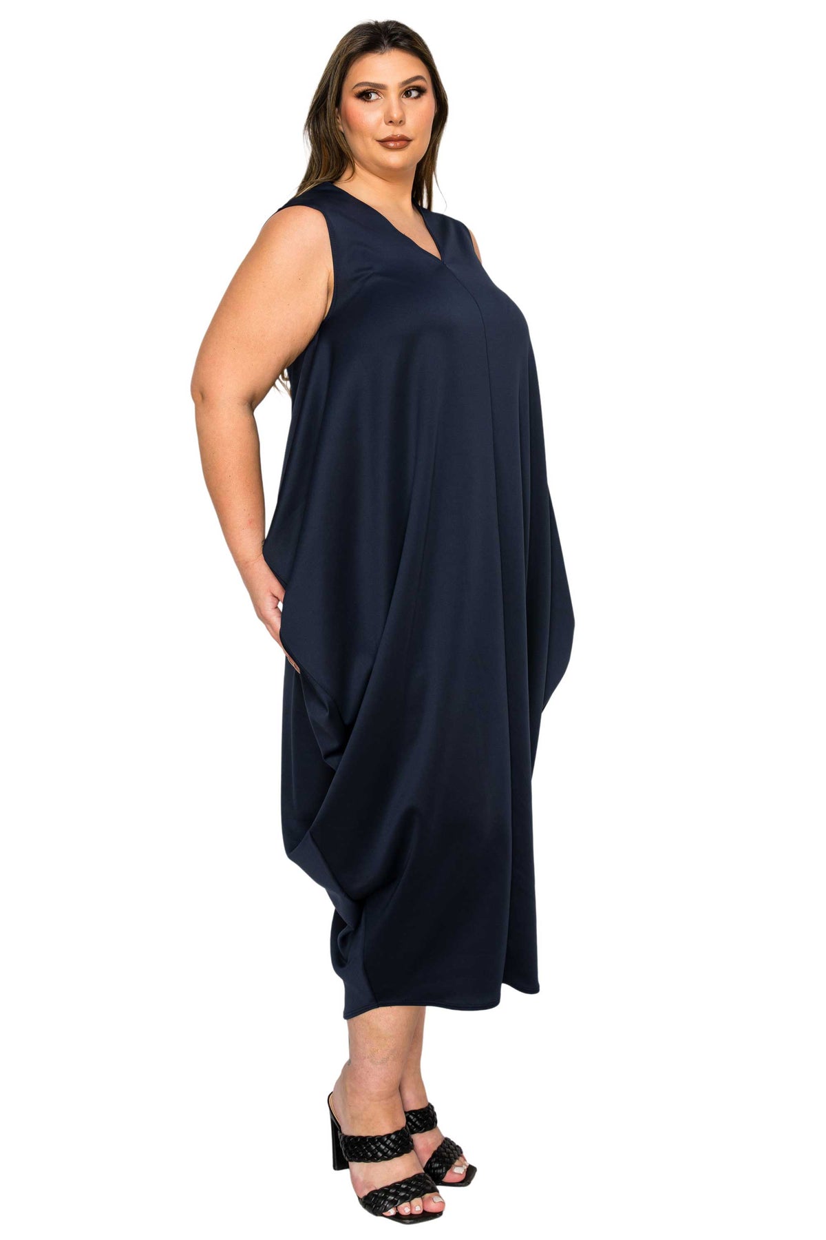 A woman modeling a sleeveless, V-neck, draped midi dress in dark navy blue, paired with black strappy heels, against a plain white background.