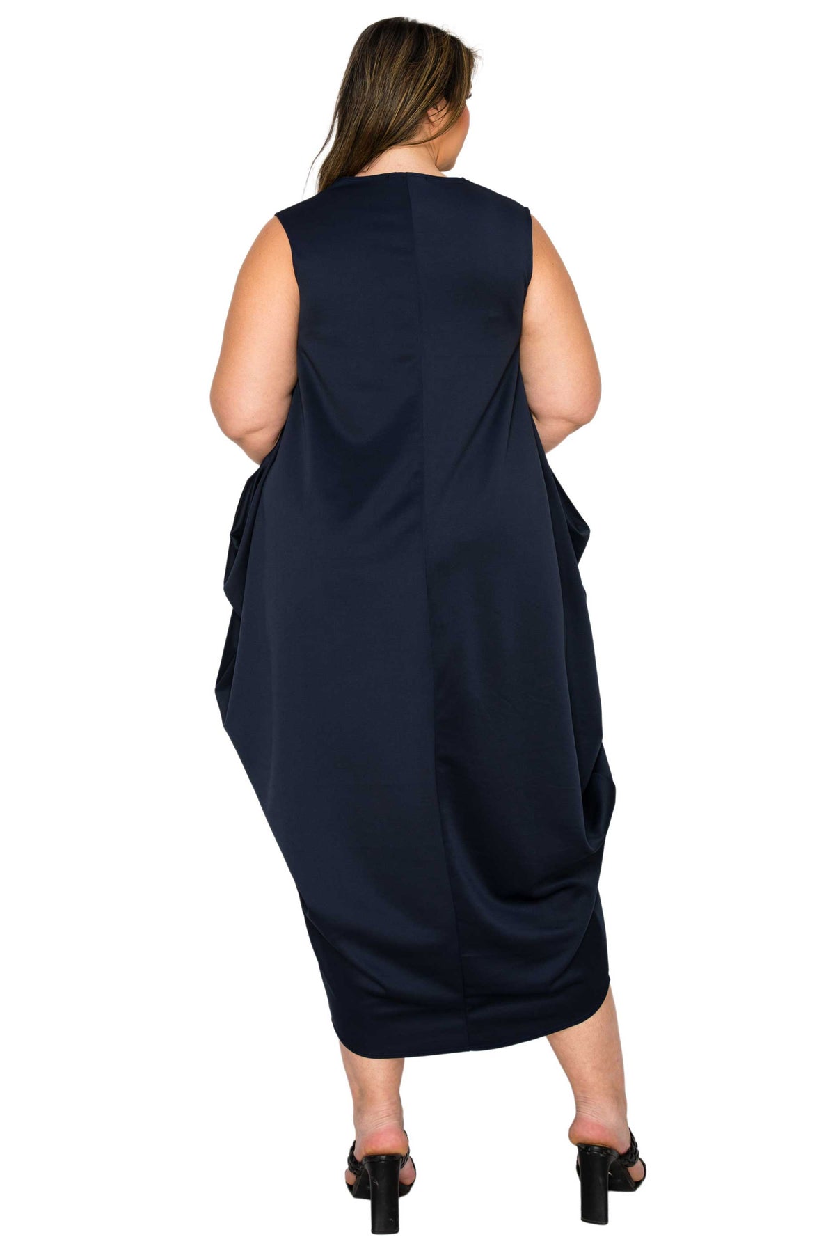 A woman viewed from the back wearing a sleeveless navy midi dress with draped side details, paired with black heeled sandals, against a plain white background.