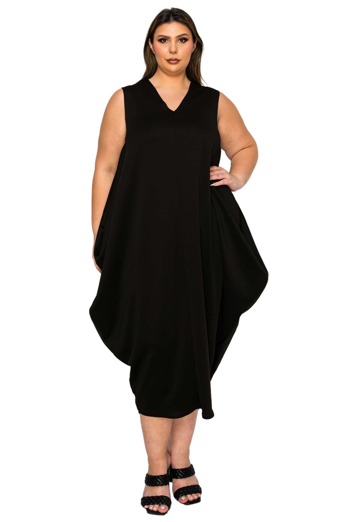 A woman modeling a sleeveless black V-neck dress with an asymmetrical draped hem, paired with black strappy heels, against a plain white background.