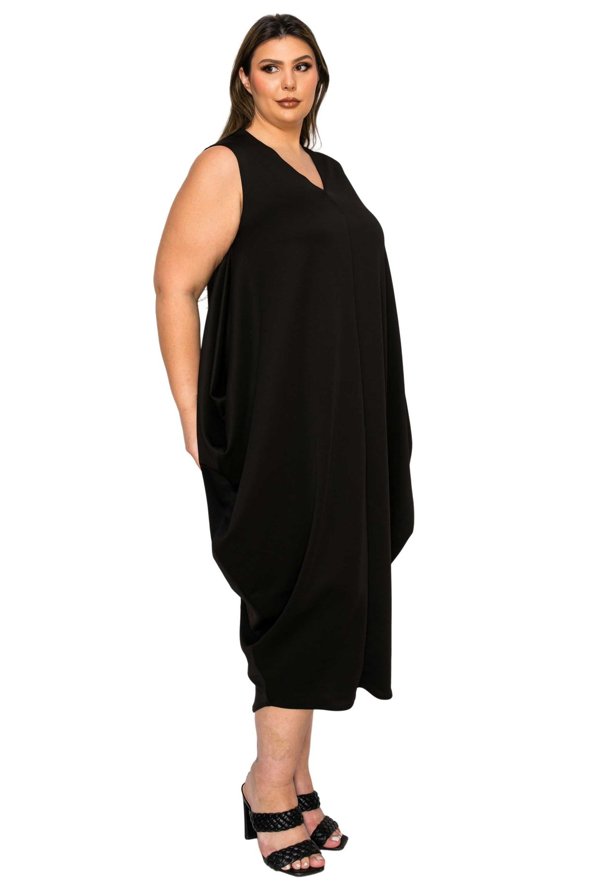 A woman modeling a sleeveless black V-neck midi dress with side pockets, paired with black strappy heels, against a plain white background.