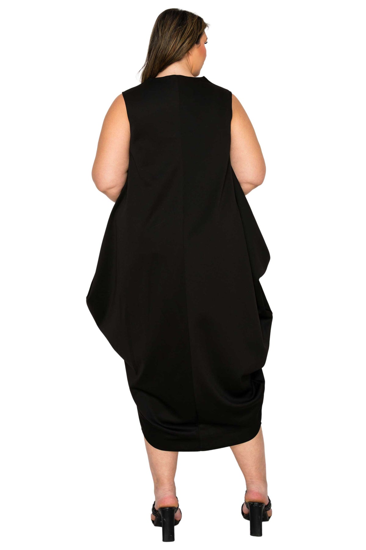 A woman viewed from the back wearing a sleeveless black dress with a draped, oversized silhouette and a gathered skirt, paired with black heels, against a plain white background.