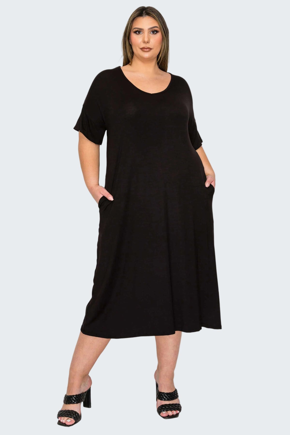 A woman modeling a black short-sleeve midi dress with side pockets, paired with black heeled sandals, against a plain white background.