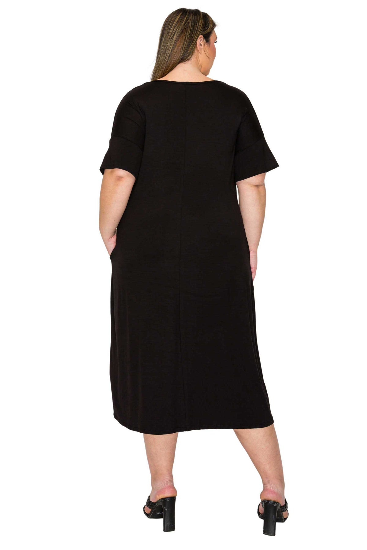 A woman viewed from the back wearing a black short-sleeve midi dress with side pockets and black heeled sandals, against a plain white studio background.