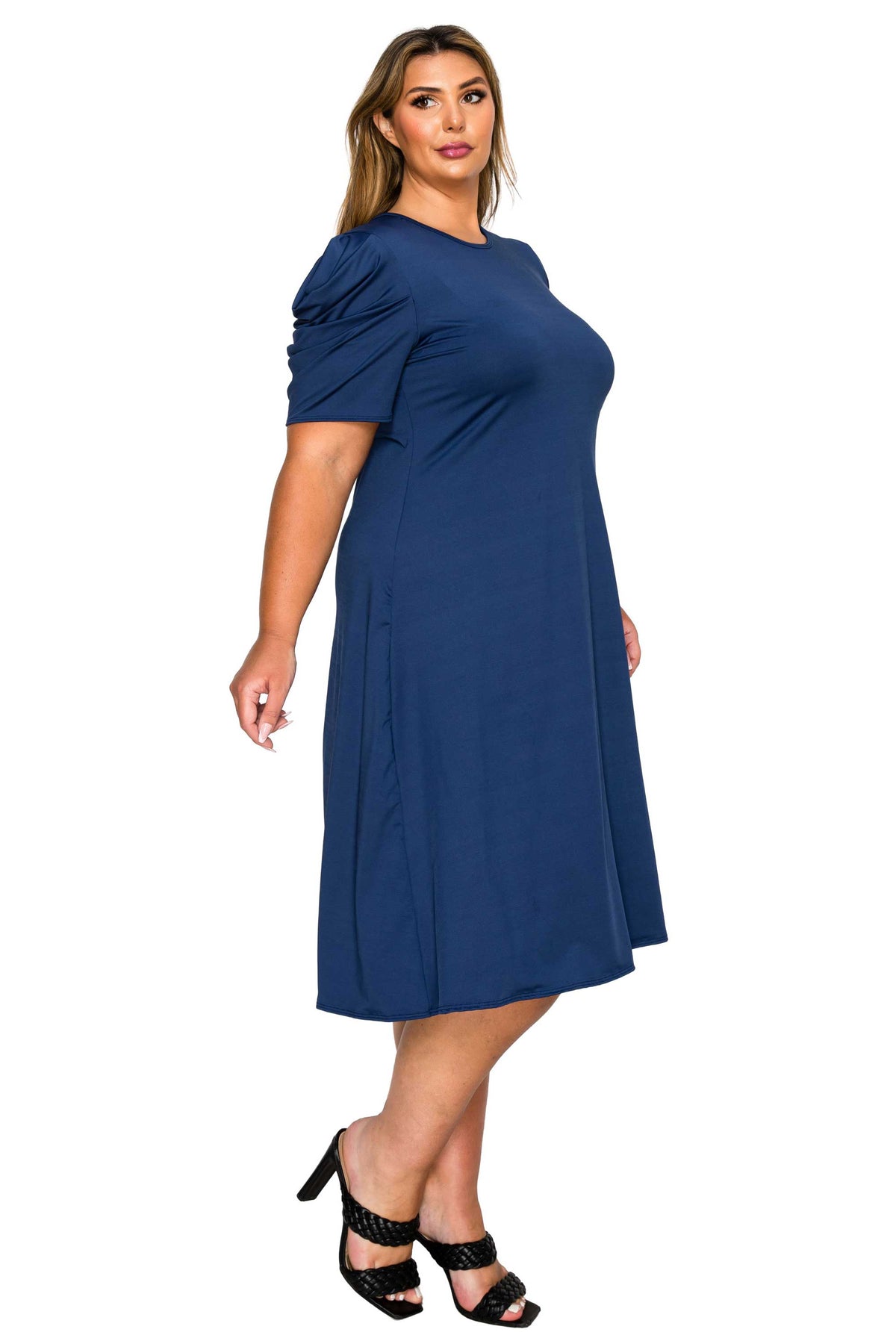 A woman modeling a navy blue midi dress with short puffed sleeves, a round neckline, and a knee-length A-line silhouette, paired with black strappy heels against a plain white background.