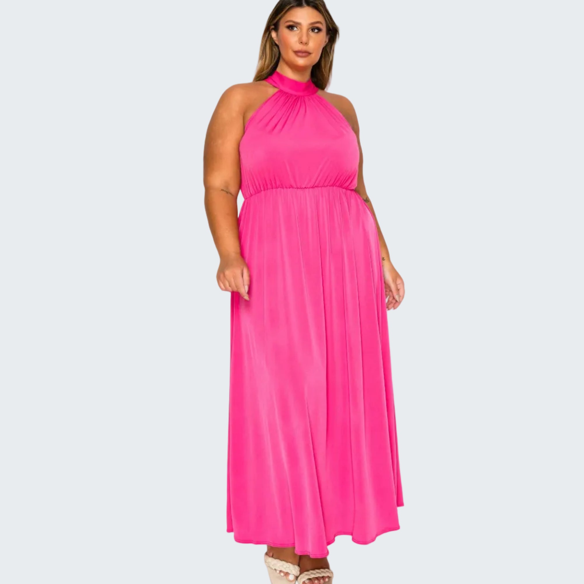 A woman modeling a bright pink halter-neck maxi dress with a gathered waist and flowing skirt, paired with beige platform sandals, against a plain white background.
