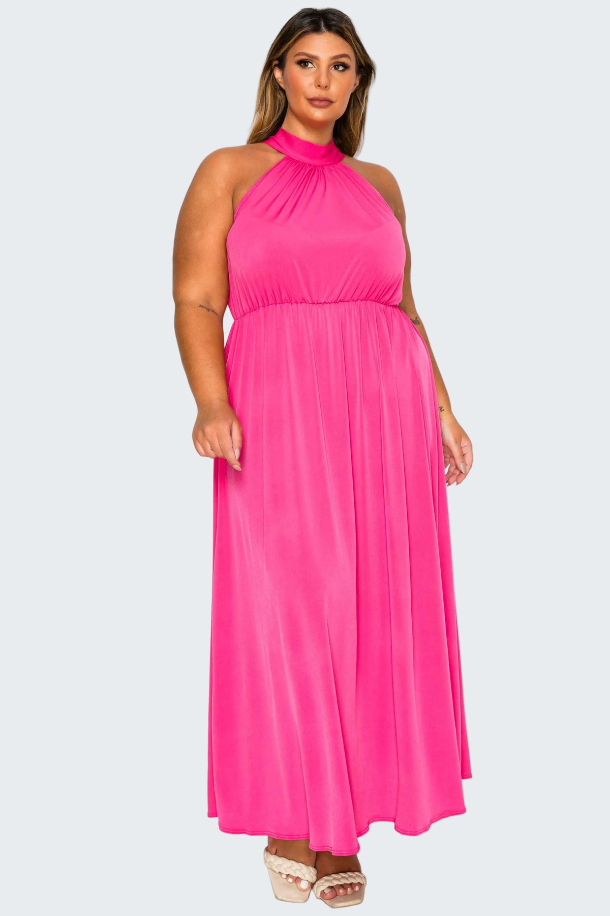 A woman modeling a bright pink halter-neck maxi dress with a gathered waist and flowing skirt, paired with beige platform sandals, against a plain white background.