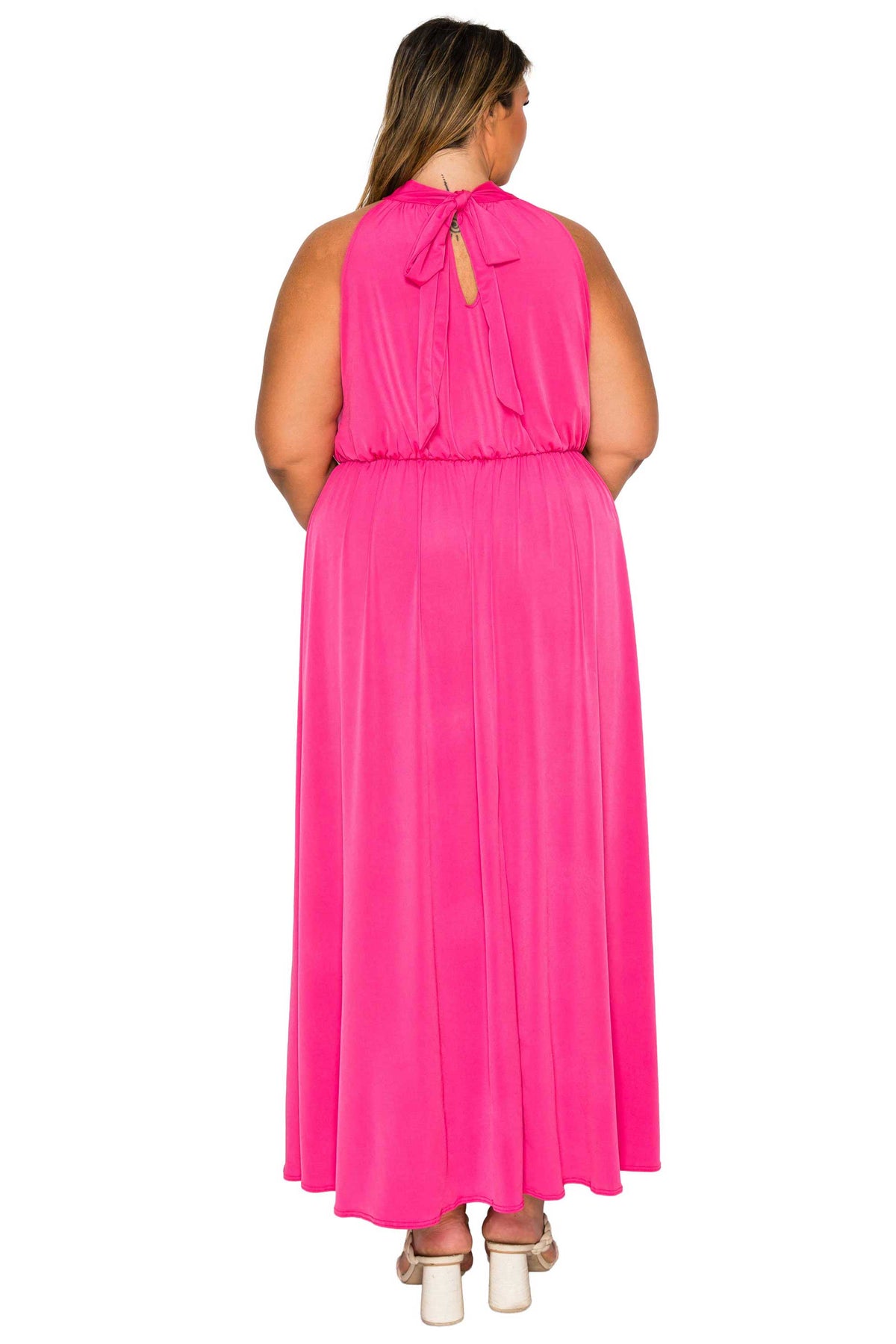 A woman viewed from the back wearing a bright pink sleeveless maxi dress with a tie-neck detail, paired with white block-heel sandals, against a plain white background.