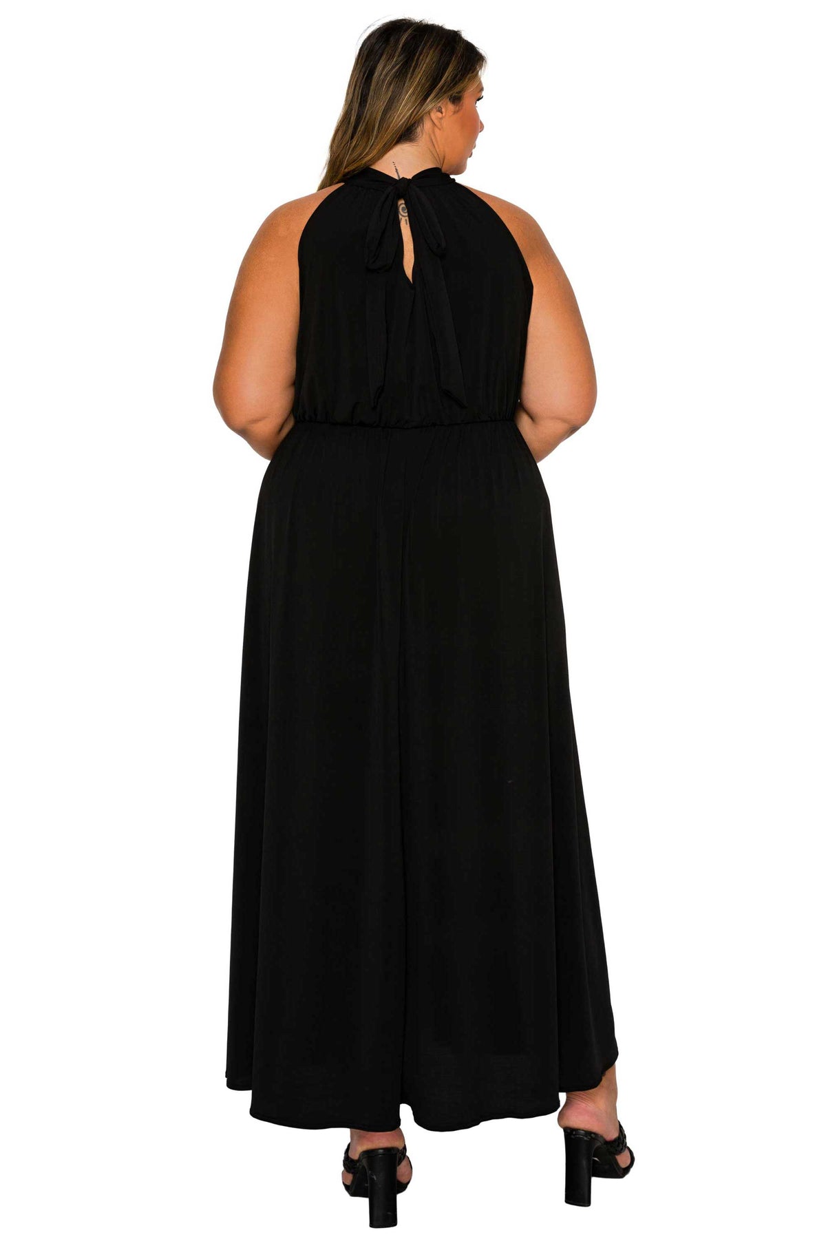 A woman viewed from the back wearing a sleeveless black maxi dress with a tie-back neckline and black heeled sandals, against a plain white studio background.