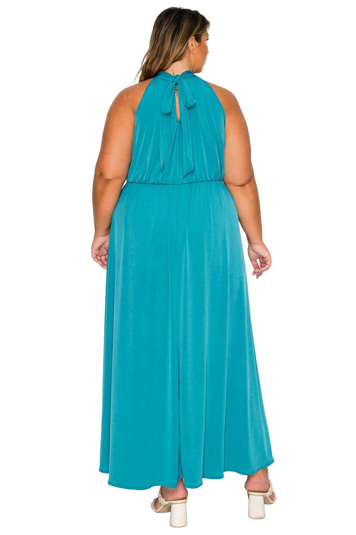 A woman viewed from the back wearing a sleeveless teal maxi dress with a tie-neck detail and flowing skirt, paired with white block-heel sandals, against a plain white background.