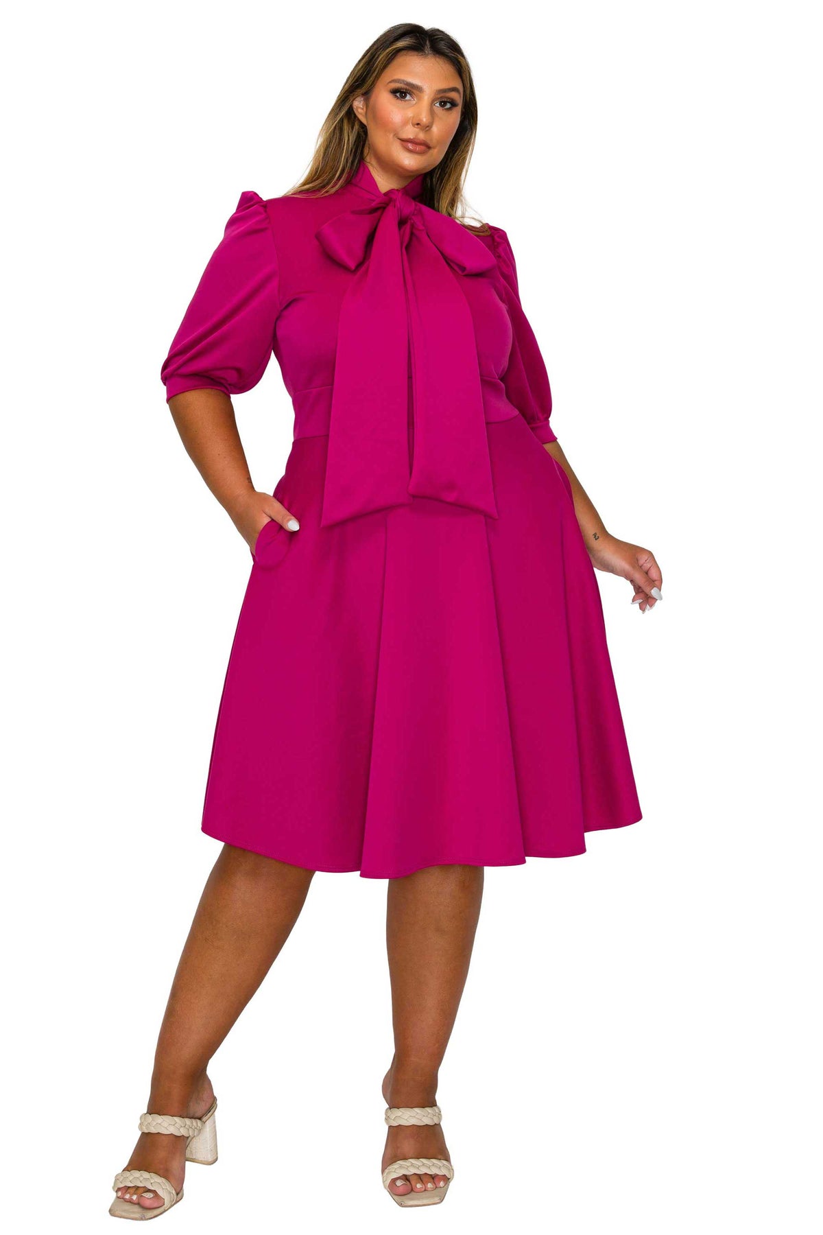 A woman modeling a bright magenta midi dress with puff sleeves, a large bow at the neckline, and a flared skirt, paired with white heeled sandals against a plain white background.