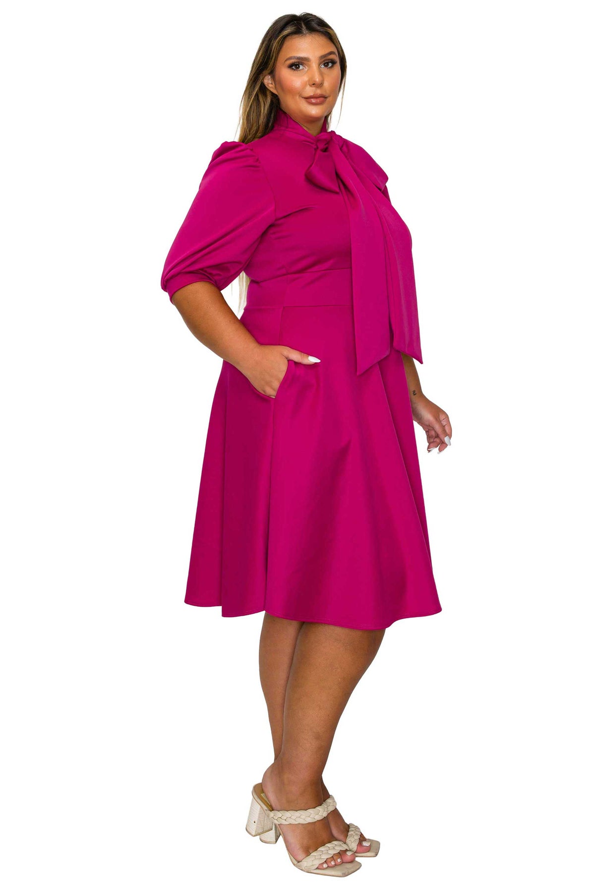 A woman modeling a bright magenta midi dress with a high neck, large bow detail at the collar, short puff sleeves, side pockets, and a flared skirt, paired with beige strappy heeled sandals against a plain white background.