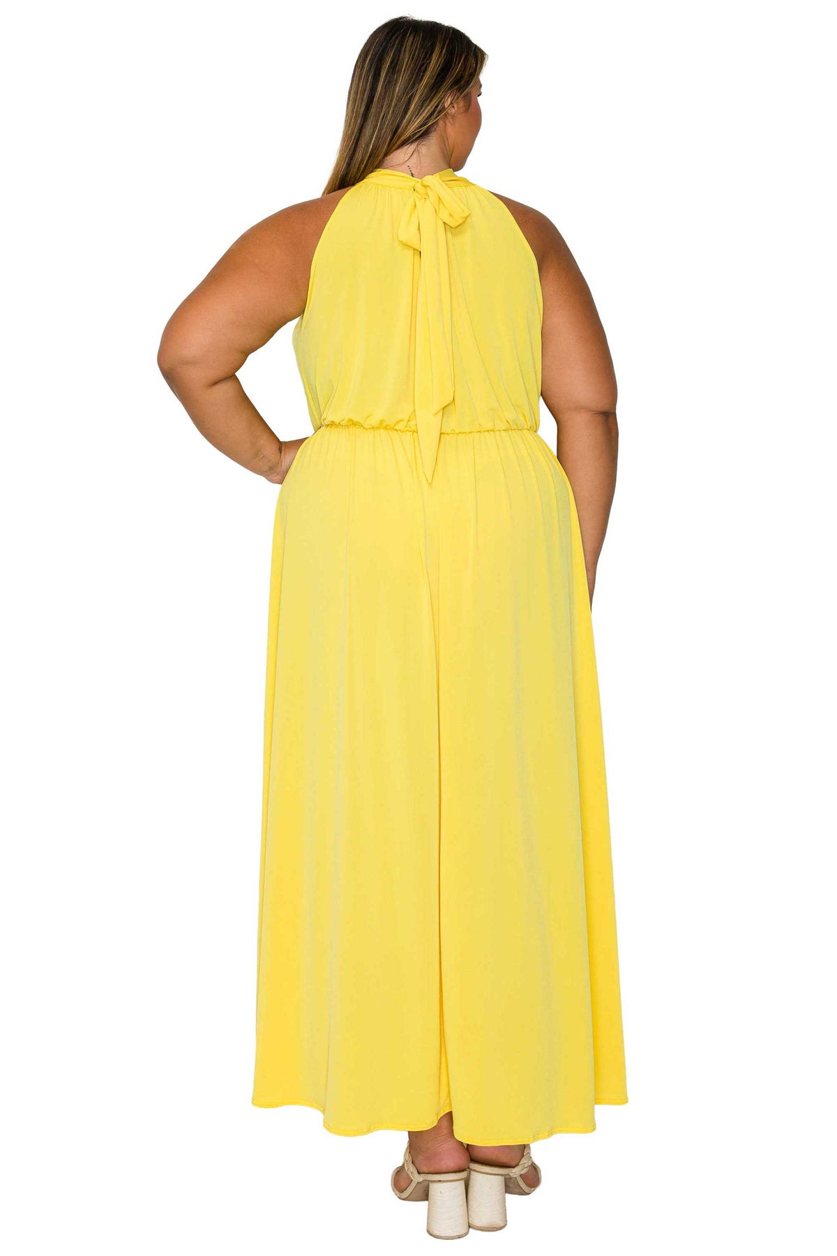 A woman shown from the back wearing a bright yellow sleeveless maxi dress with a tie-neck detail and a flared skirt, paired with white block-heel sandals, against a plain white background.