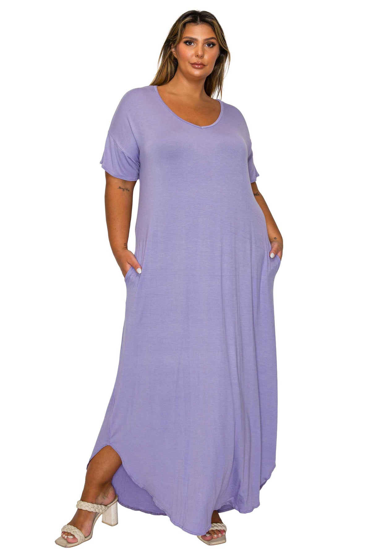 A woman modeling a loose, short-sleeve maxi dress in a light lavender purple color, featuring side pockets and a high-low hem, paired with white heeled sandals against a plain white background.