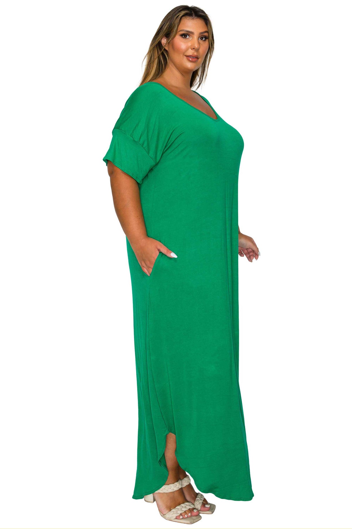 A woman modeling a bright green, short-sleeved maxi dress with a V-neckline and side pockets, paired with white heeled sandals, set against a plain white background.