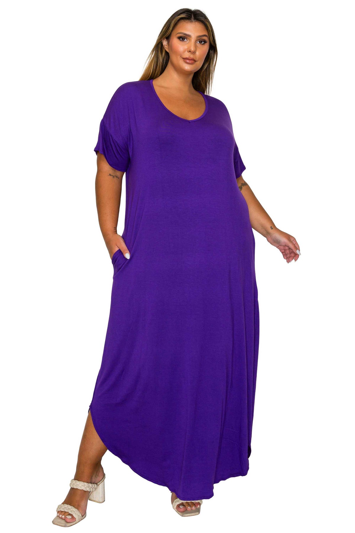 A woman modeling a loose-fitting, short-sleeve maxi dress in solid purple with side pockets, paired with white heeled sandals, against a plain white background.