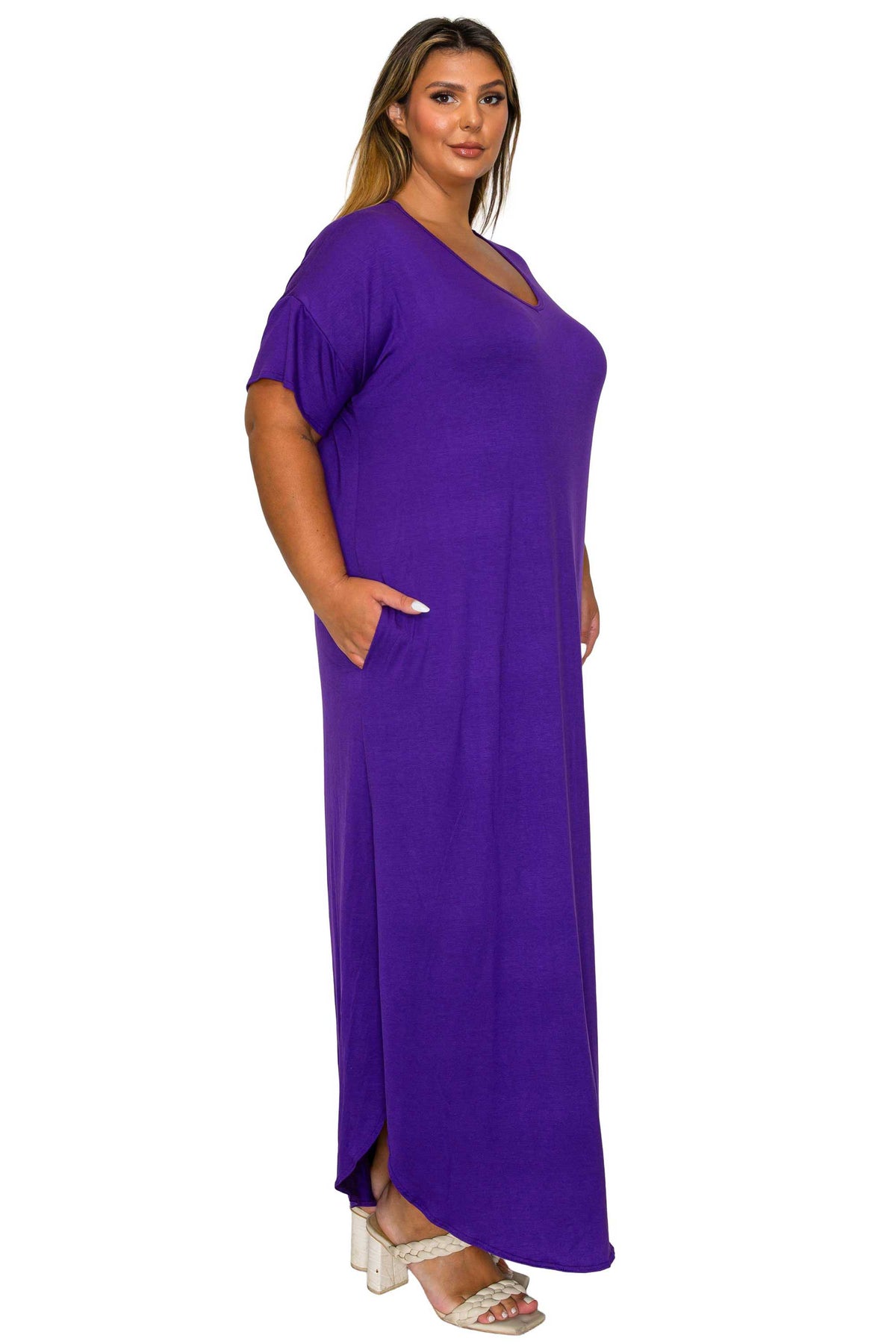 A woman modeling a purple short-sleeve maxi dress with a V-neckline and side pockets, paired with white heeled sandals, against a plain white background.
