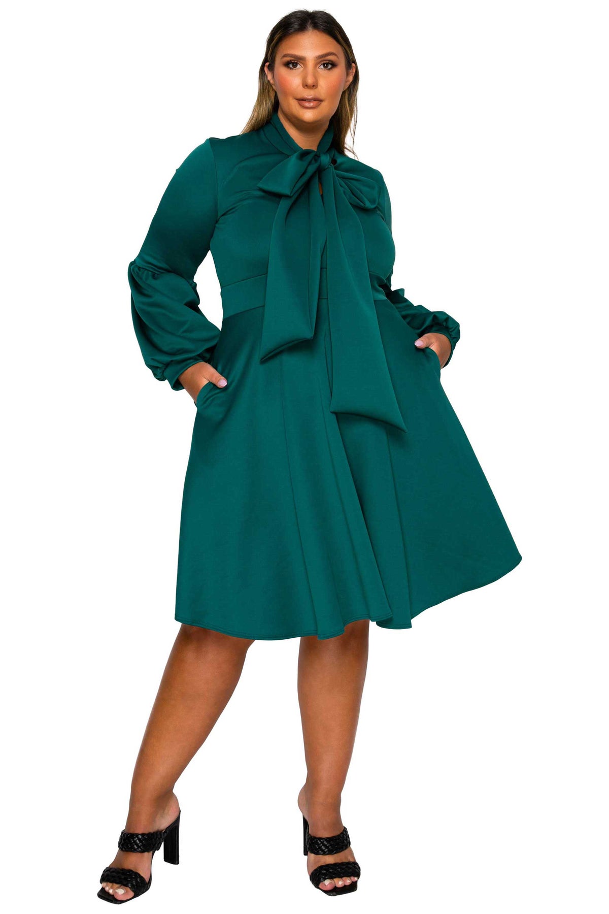 A woman modeling a teal green long-sleeve midi dress with a large bow at the collar, side pockets, and a flared skirt, paired with black heeled sandals against a plain white background.