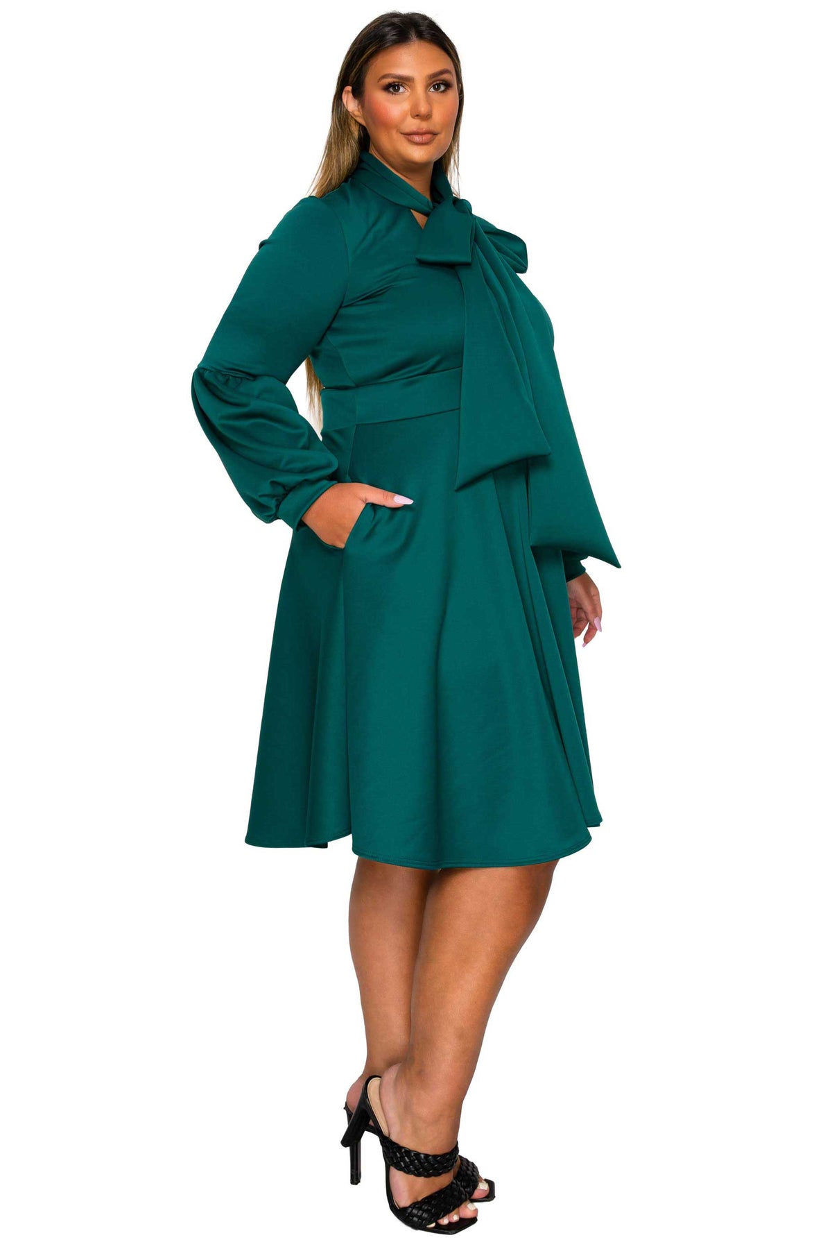A woman modeling a teal green long-sleeve dress with a high ruffled collar, tie detail at the neckline, and a flared skirt, paired with black strappy heels against a plain white background.