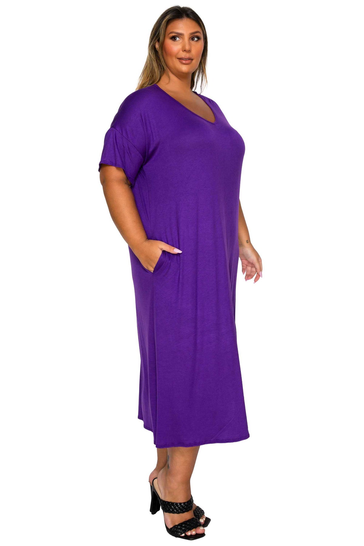 A woman modeling a purple short-sleeve V-neck midi dress with side pockets, paired with black strappy heels, against a plain white background.