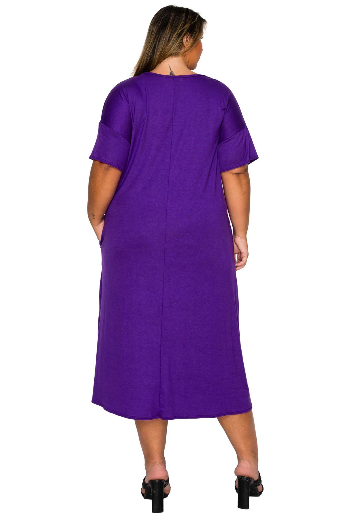 A woman viewed from the back wearing a solid purple midi dress with short sleeves and black heeled sandals, set against a plain white background.