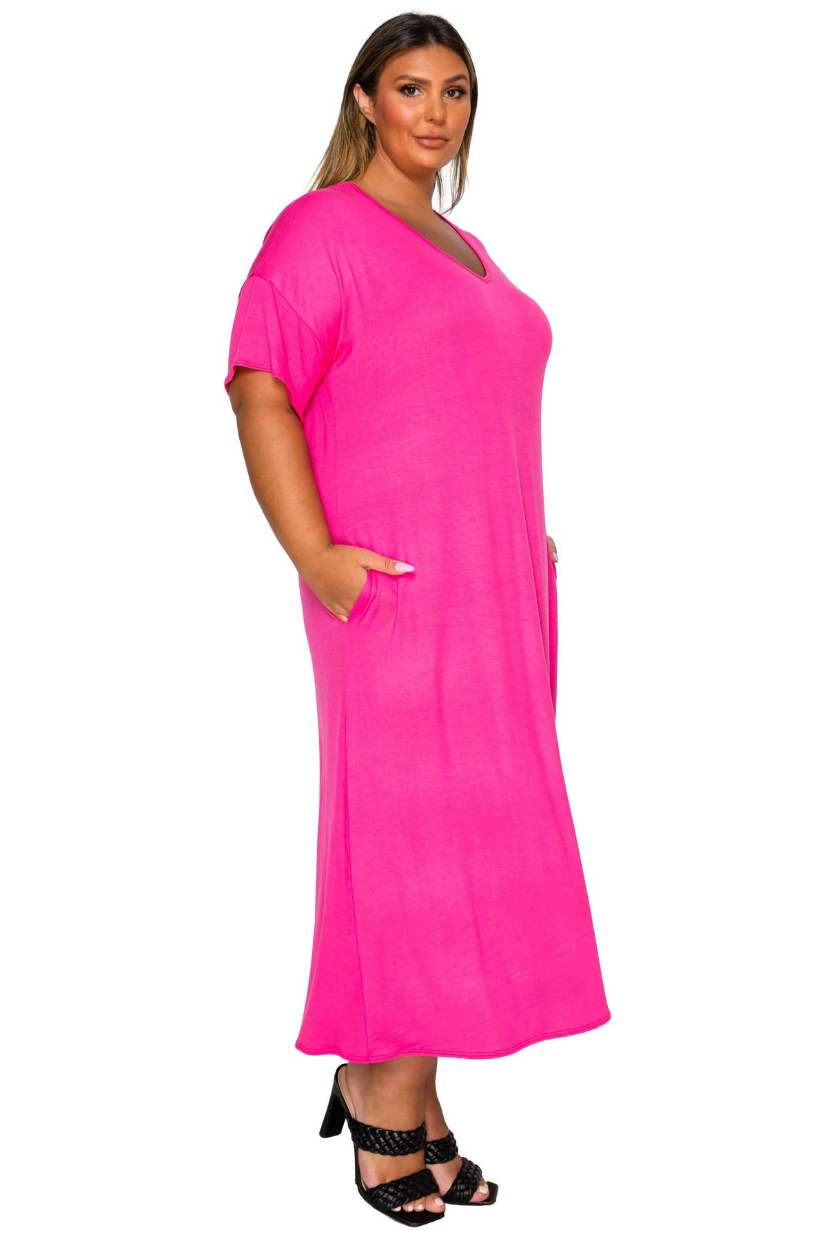 A woman modeling a bright pink short-sleeve maxi dress with side pockets, paired with black strappy heels, against a plain white background.