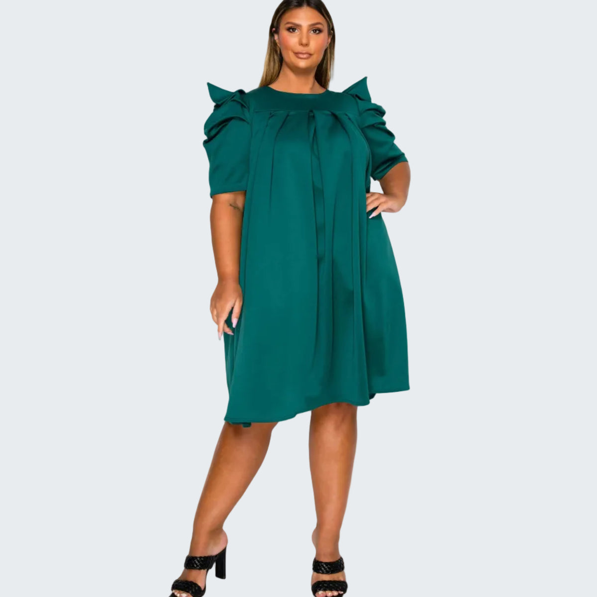 A woman modeling a teal green shift dress with dramatic ruffled puff sleeves and a tie-neck detail, paired with black heeled sandals against a plain white background.