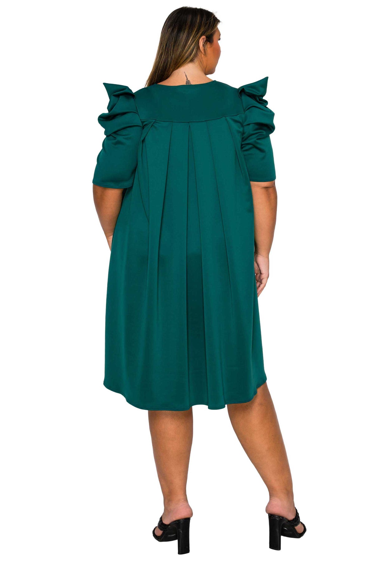A woman viewed from the back wearing a dark teal green dress with dramatic ruffled puff sleeves and a pleated bodice, paired with black block-heel mules, against a plain white background.