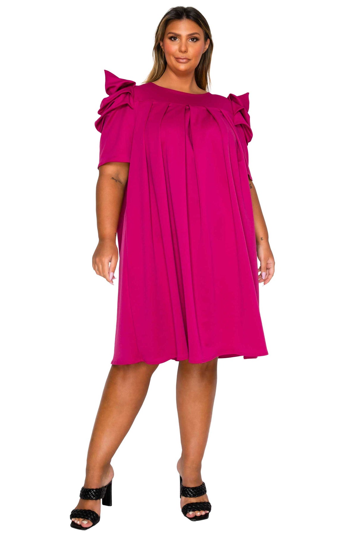 A woman modeling a bright fuchsia pink short-sleeve dress with dramatic ruffled shoulder details and a gathered waist, paired with black heeled sandals against a plain white background.