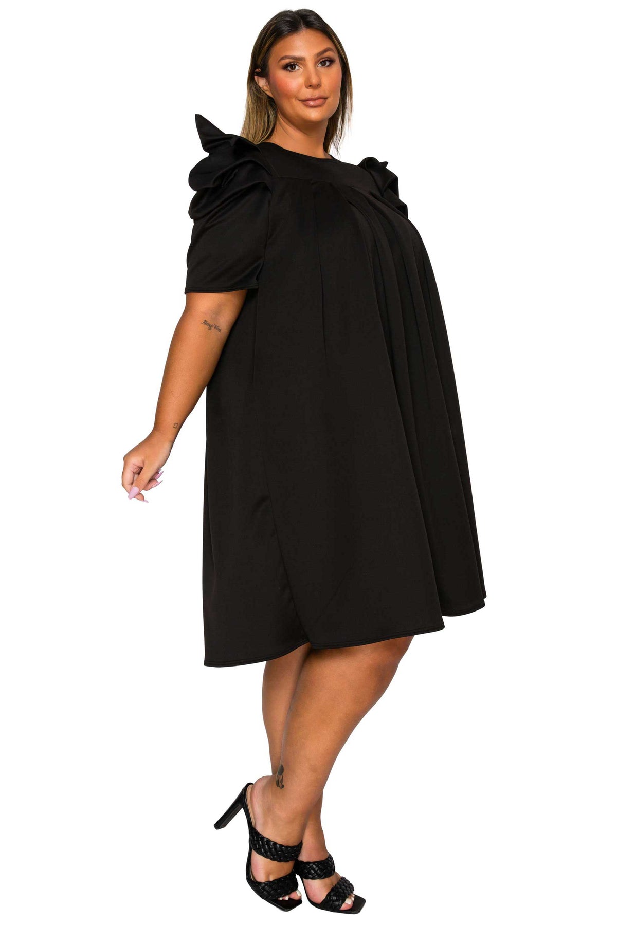 A woman modeling a black short-sleeve dress with dramatic ruffled shoulder details, photographed against a plain white studio background.