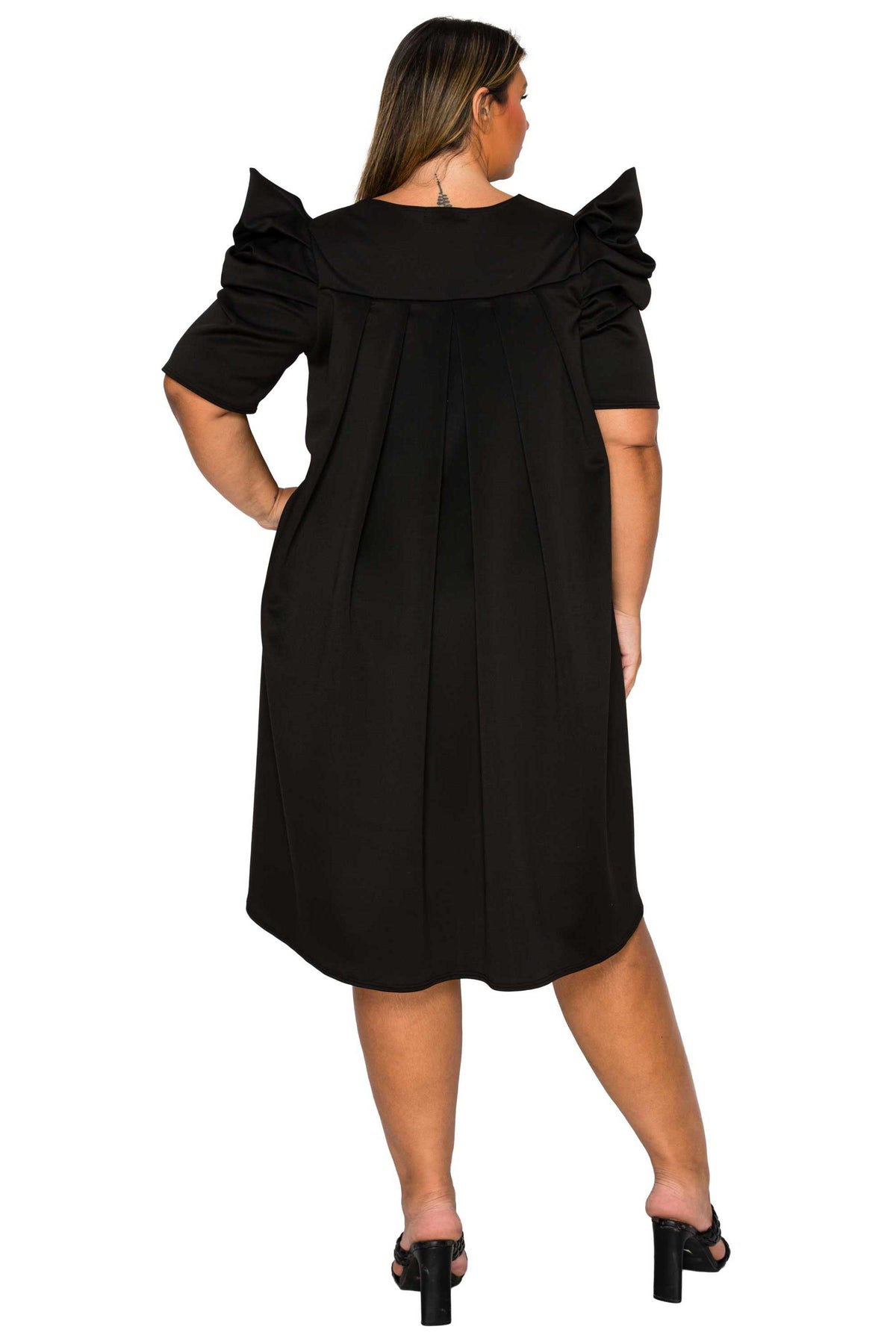A woman modeling a black short-sleeve dress from the back, featuring dramatic ruffled shoulder details and a pleated skirt, set against a plain white background.