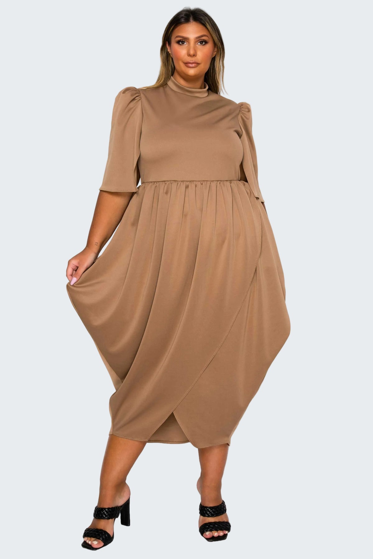 A woman modeling a taupe brown midi dress with puff sleeves, a mock neck, and an asymmetrical draped hem, paired with black heeled sandals against a plain white background.
