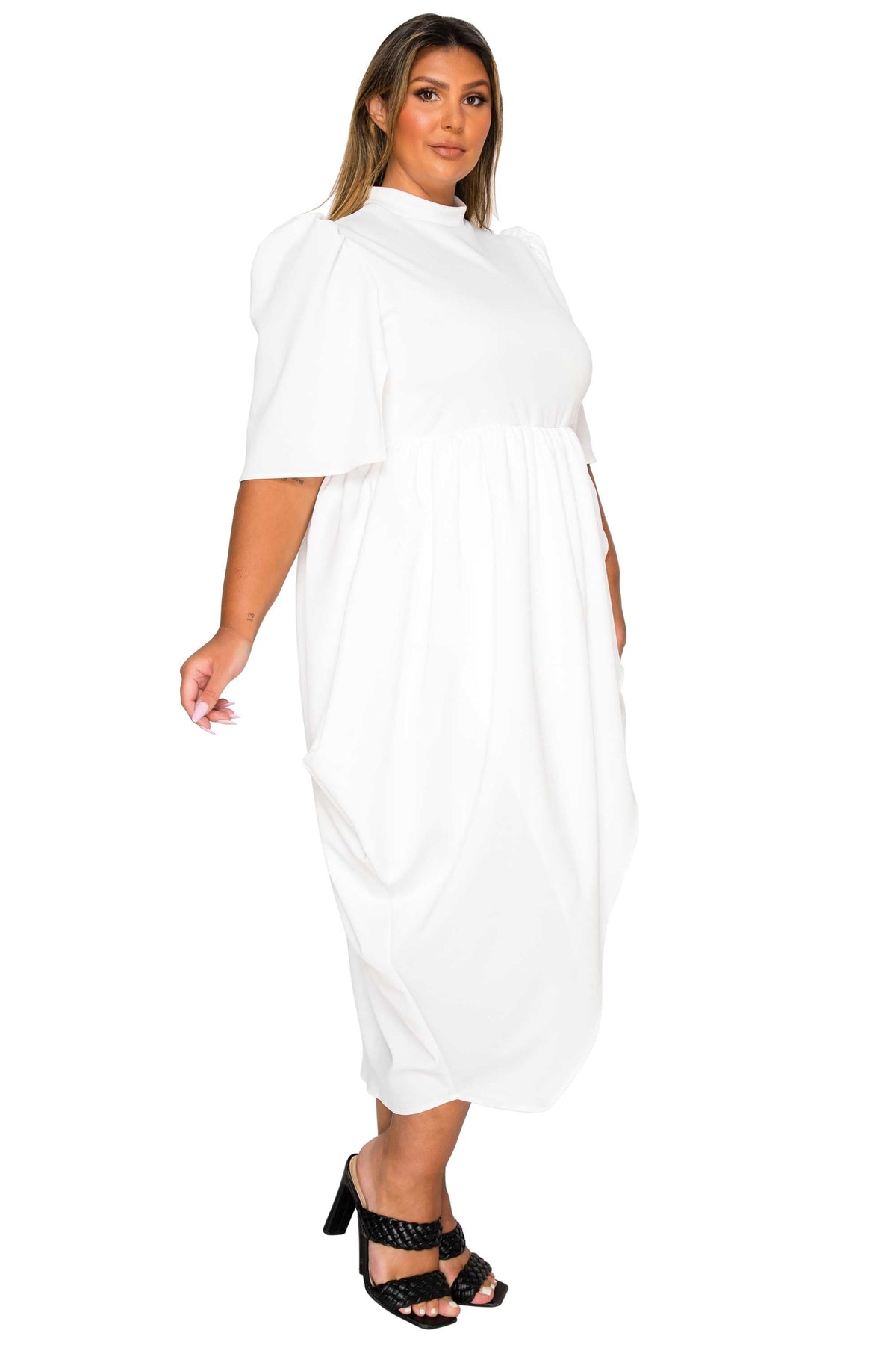A woman modeling a white midi dress with short puff sleeves, a high mock neck, and a gathered waist, paired with black strappy heels against a plain white background.