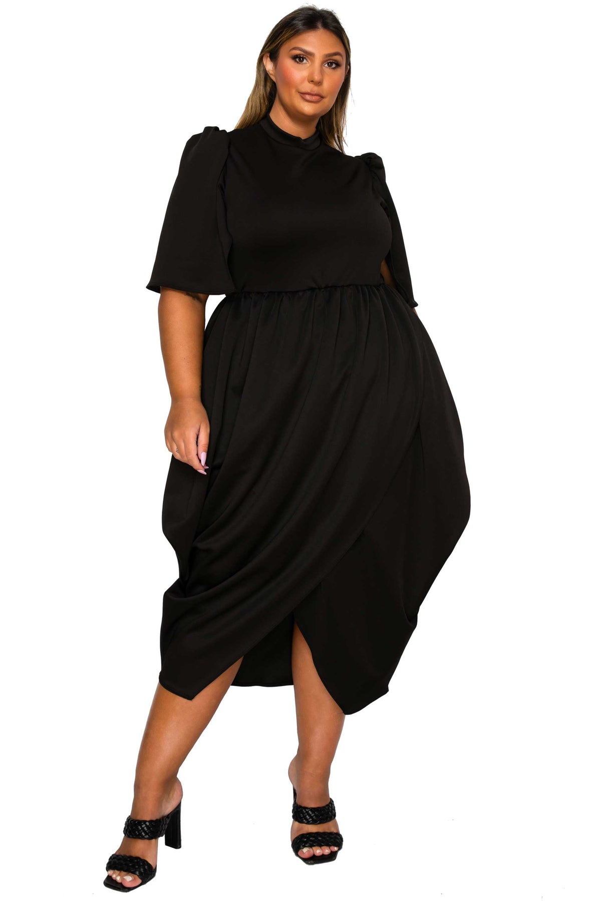 A woman modeling a black midi dress with short puff sleeves, a high mock neck, and a draped wrap-style skirt, paired with black heeled sandals against a plain white background.