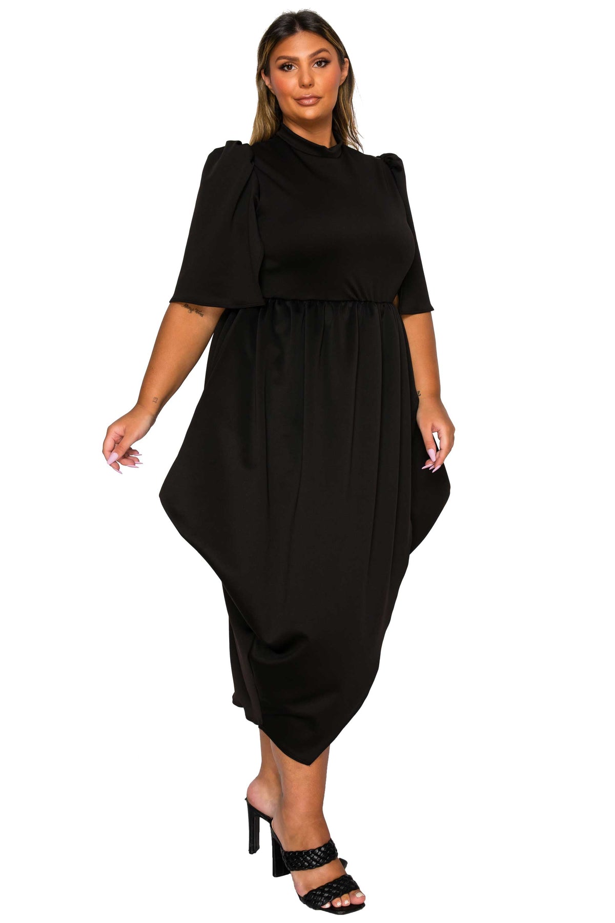 A woman modeling a black midi dress with short puff sleeves, a high mock neck, and an asymmetrical draped hem, paired with black heeled sandals against a plain white background.