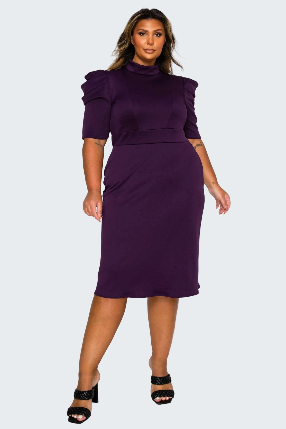 A woman modeling a deep purple midi dress with puffed short sleeves, a mock neck, and a fitted waist, paired with black heeled sandals against a plain white background.