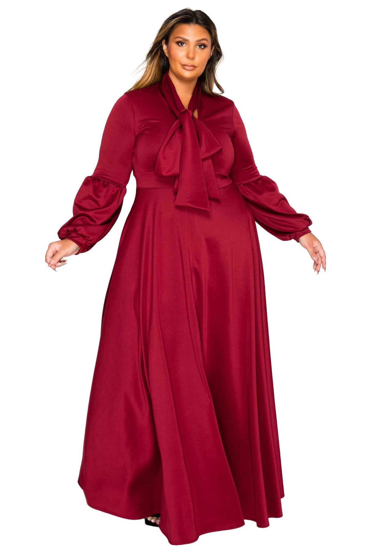 A woman modeling a long-sleeved, floor-length burgundy dress with a large bow tie-neck detail, photographed against a solid white background.