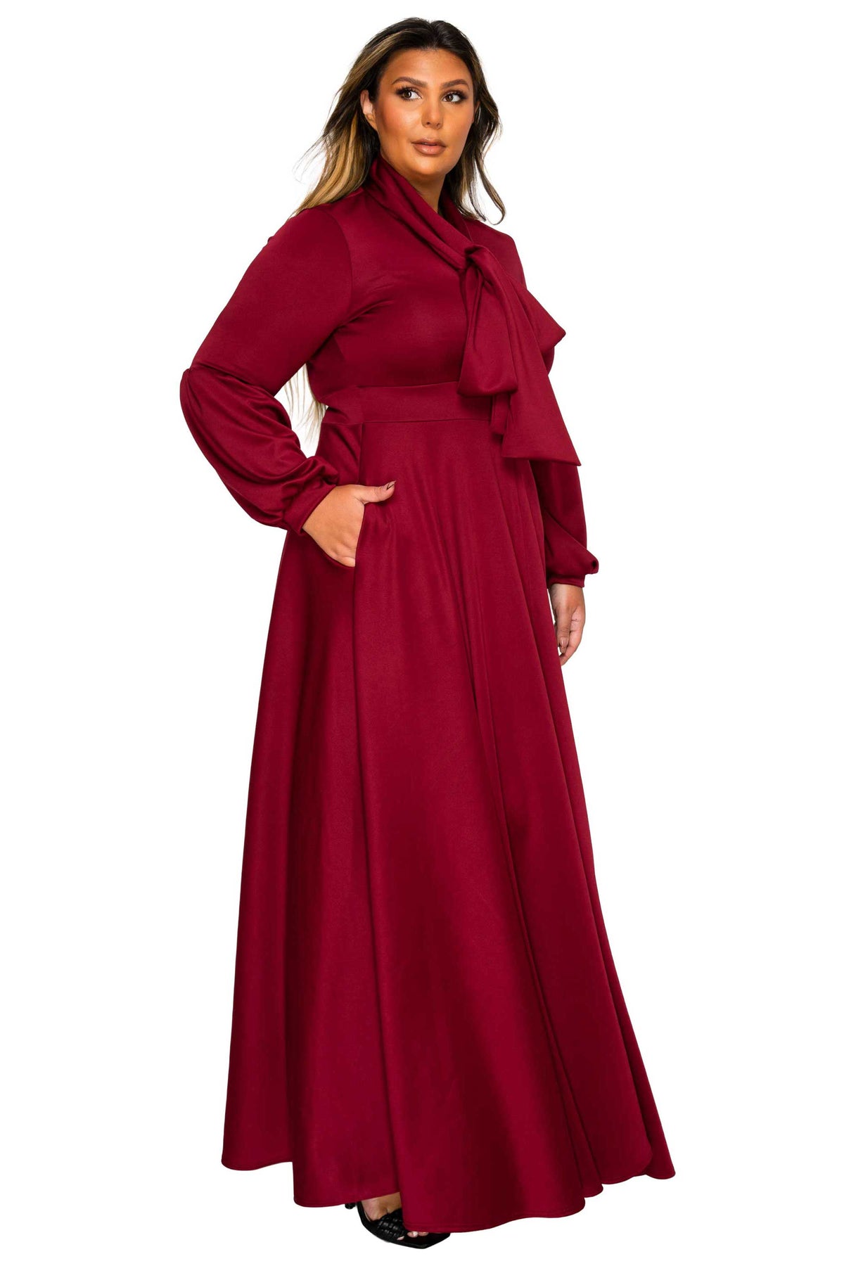A woman modeling a long-sleeved, floor-length magenta maxi dress with a ruffled collar, waist tie, and side pockets, set against a plain white background.