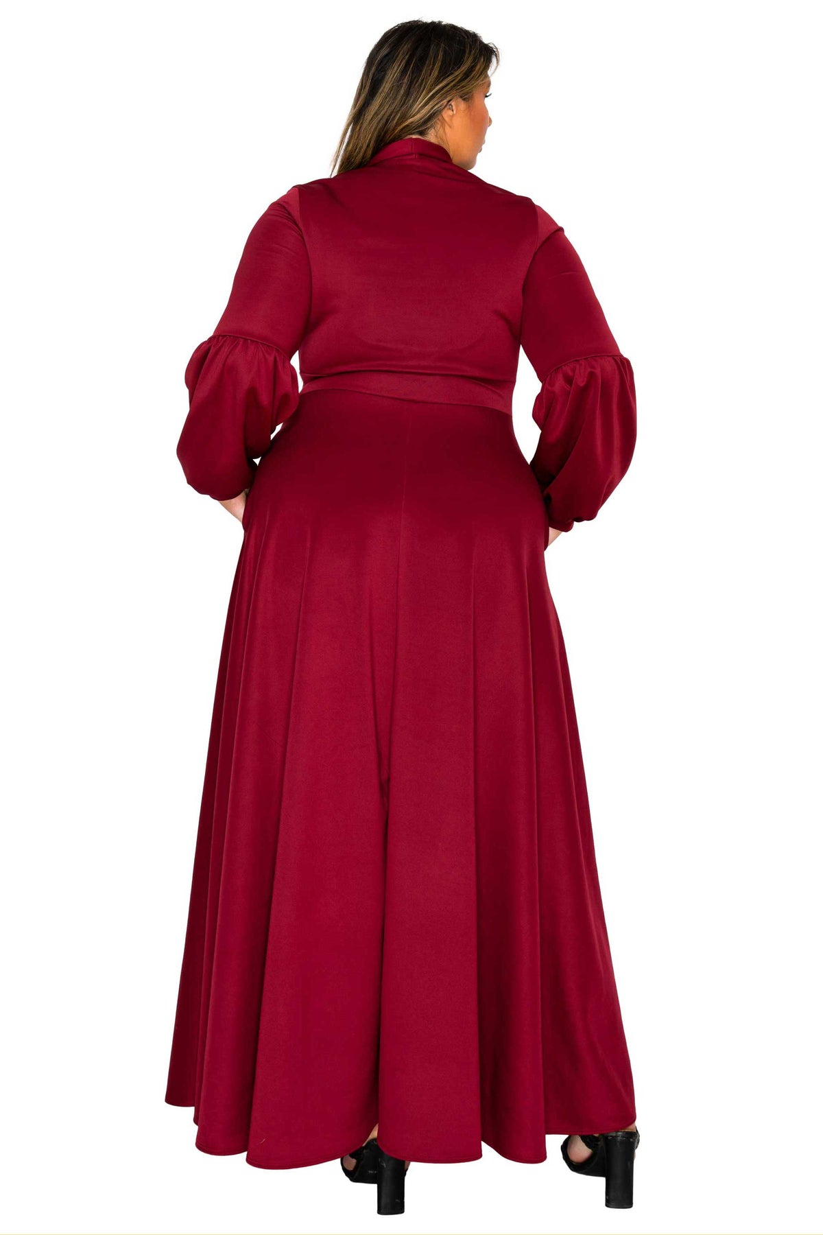 A woman viewed from the back wearing a long, flowing burgundy maxi dress with long puffed sleeves and a high collar, paired with black heels, against a plain white background.