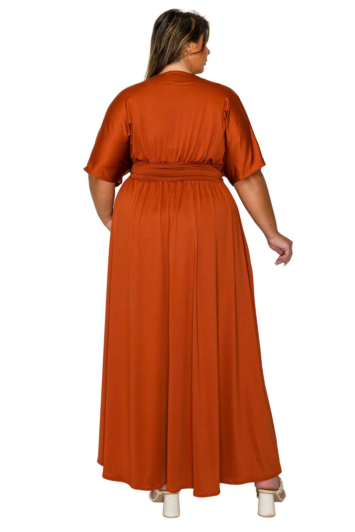 A woman viewed from the back wearing a rust-orange maxi dress with short sleeves and a gathered waist, paired with white heeled sandals against a plain white background.