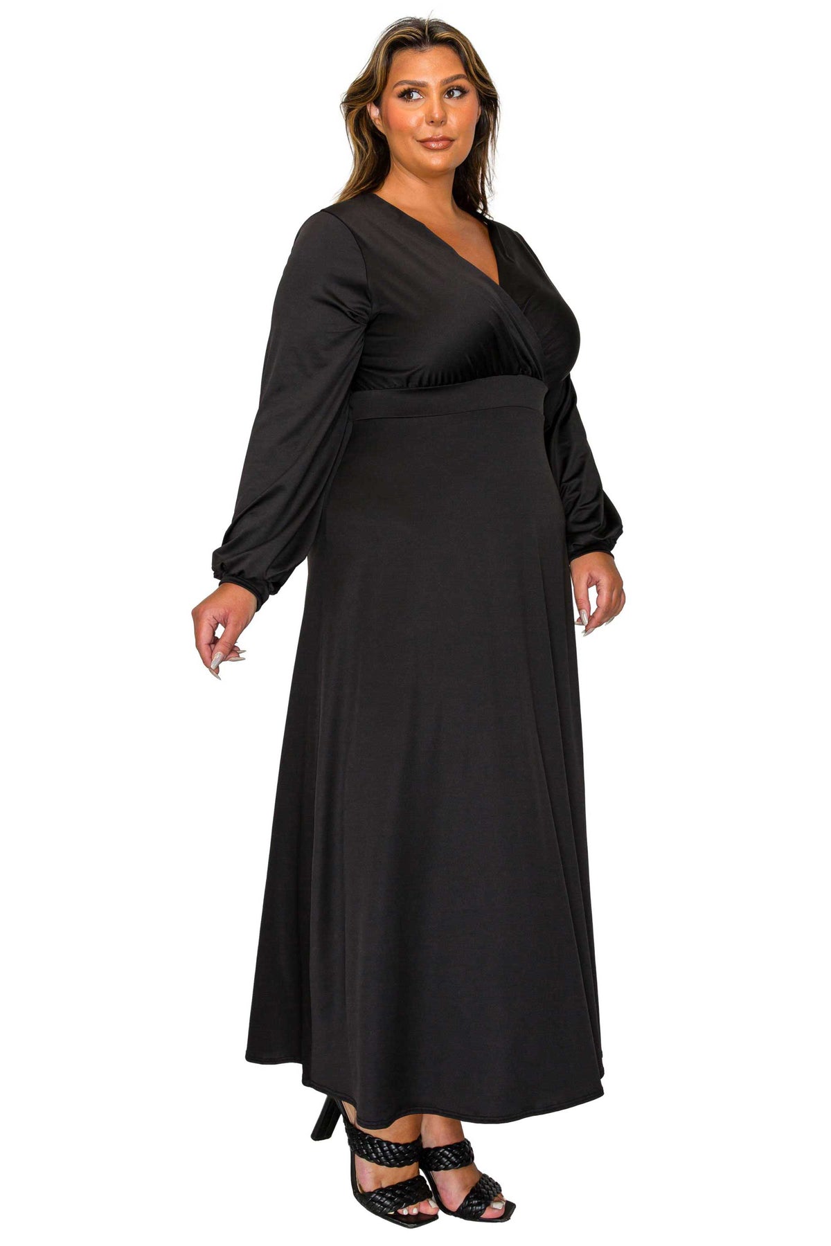 A woman modeling a long black wrap-style maxi dress with long sleeves and black strappy heels against a plain white background.
