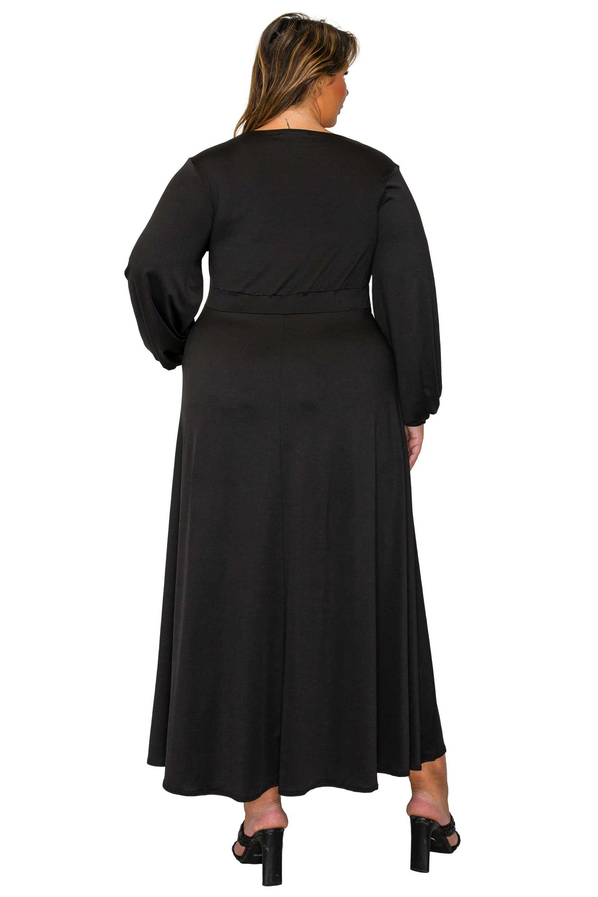 A woman viewed from the back wearing a long black dress with long sleeves and a matching waistband, paired with black high-heeled sandals, against a plain white background.