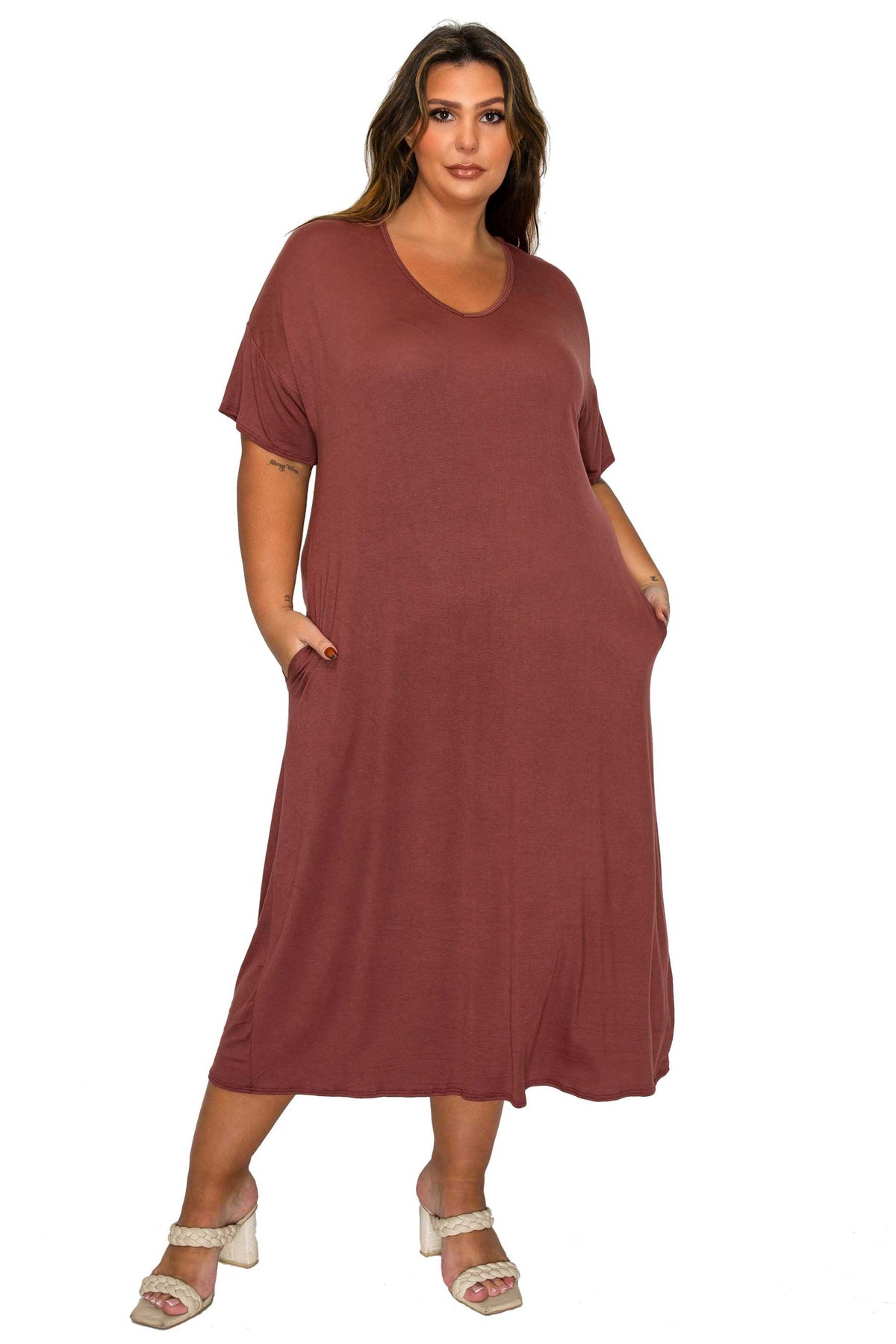A woman modeling a loose-fitting, short-sleeve maxi dress in a muted terracotta or dusty rose-brown color, featuring side pockets and a relaxed drape, paired with white heeled sandals against a plain white background.