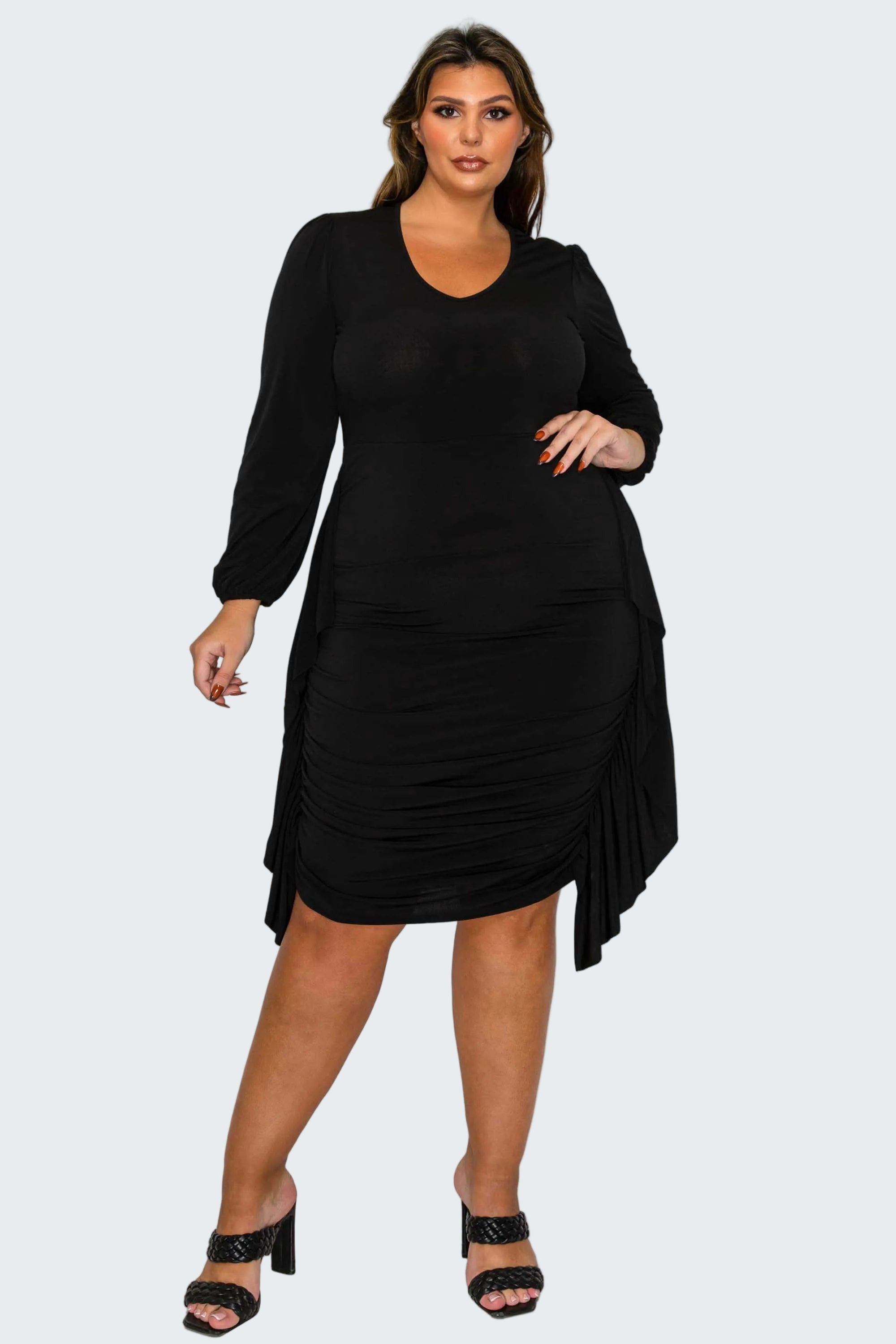 A woman modeling a black long-sleeve midi dress with ruched detailing and an asymmetrical draped hem, paired with black heeled sandals, against a plain white background.