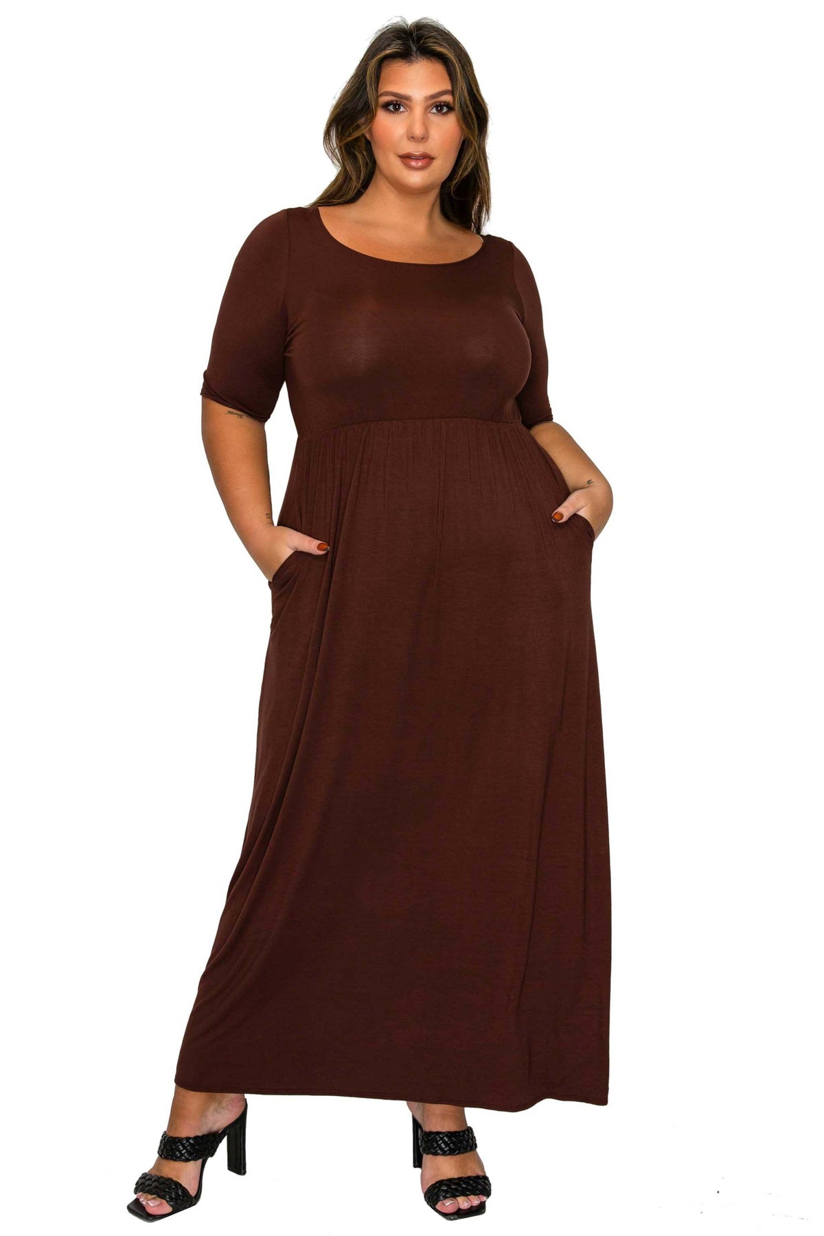 A woman modeling a dark brown short-sleeve maxi dress with side pockets, paired with black heeled sandals, against a plain white background.