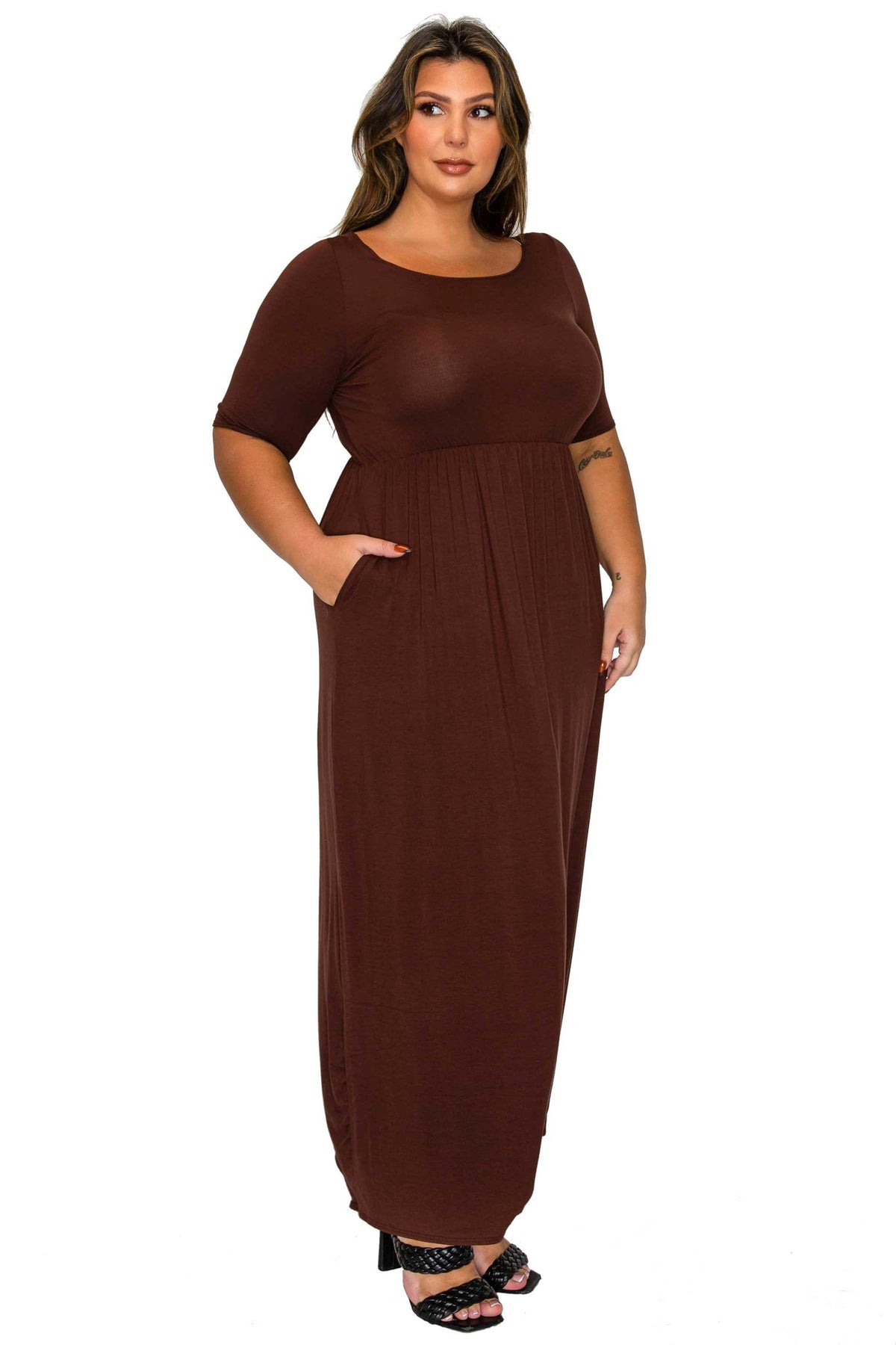 A woman modeling a brown short-sleeve maxi dress with a relaxed, gathered skirt and side pockets, paired with black heeled sandals, against a plain white background.