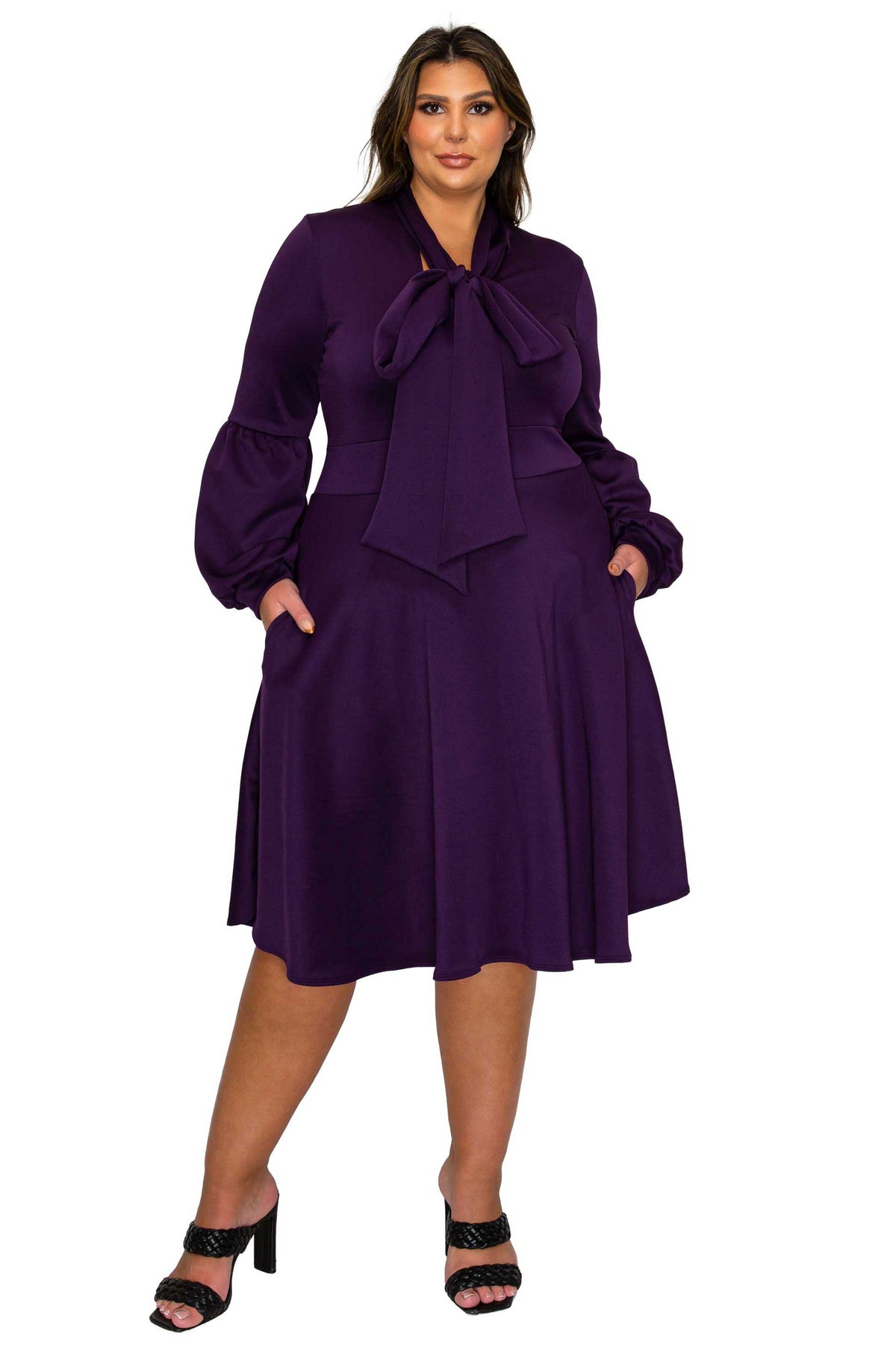 A woman modeling a knee-length purple dress with long balloon sleeves, a large bow at the neckline, and side pockets, paired with black heeled sandals against a plain white background.