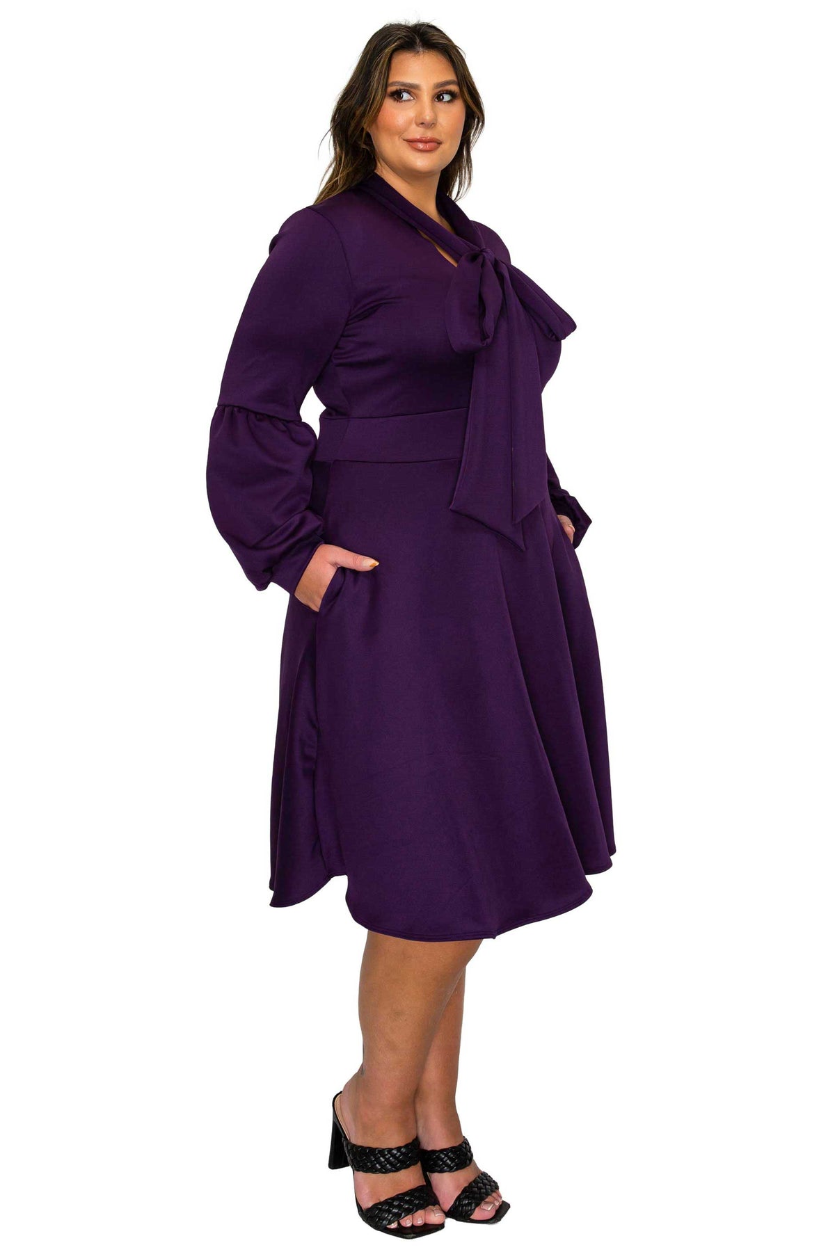 A woman modeling a knee-length purple wrap dress with long balloon sleeves, a large bow at the neckline, side pockets, and a matching waist tie, paired with black heeled sandals against a plain white background.