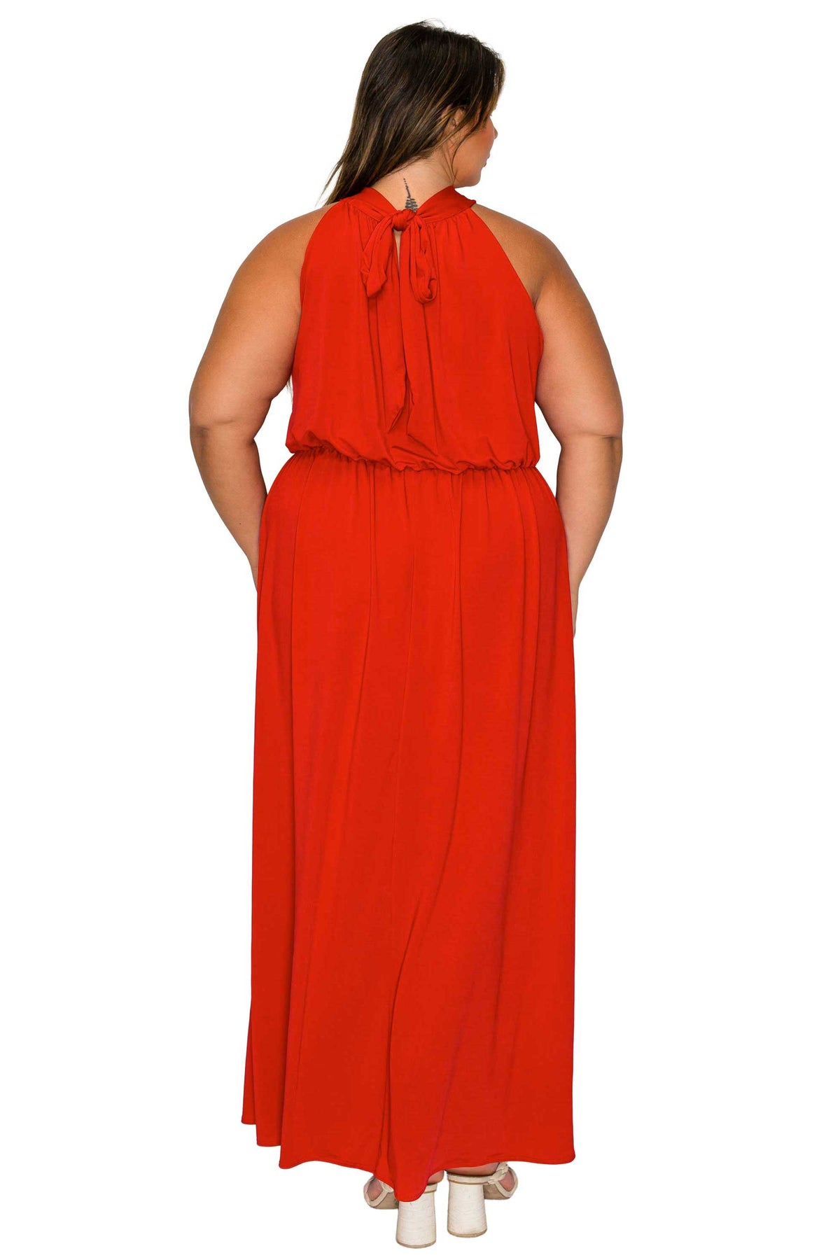 A woman viewed from the back wearing a bright red sleeveless maxi dress with a tie-neck detail and gathered waist, paired with white heels against a plain white background.