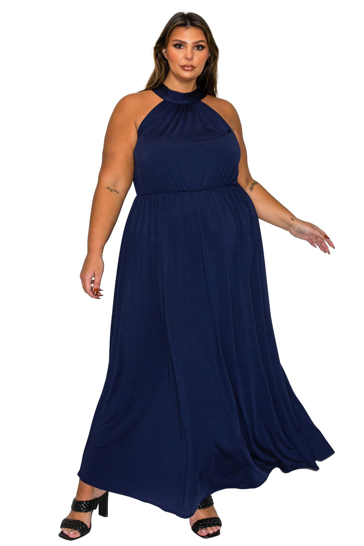 A woman modeling a navy blue halter-neck maxi dress with a gathered waist and flowing skirt, paired with black heeled sandals, against a plain white background.